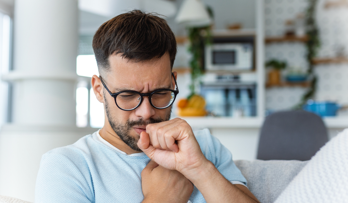 Man wearing glasses sitting on a couch, coughing into his fist, with a concerned expression, in a modern and brightly lit living room.