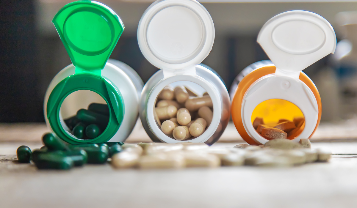 Three open bottles of multivitamins in vibrant green, white, and orange caps, displaying a variety of capsules, tablets, and soft gels, placed on a wooden surface with a blurred background.